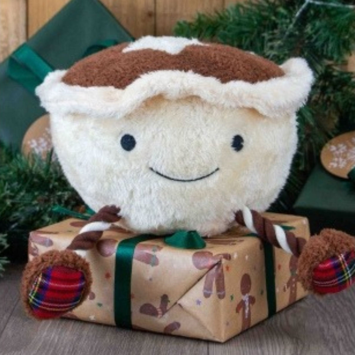 A plush mince pie character dog toy with a cream-colored body, brown crust, and embroidered smiley face, sitting on top of a wrapped Christmas gift box with green ribbon and holiday decorations in the background.