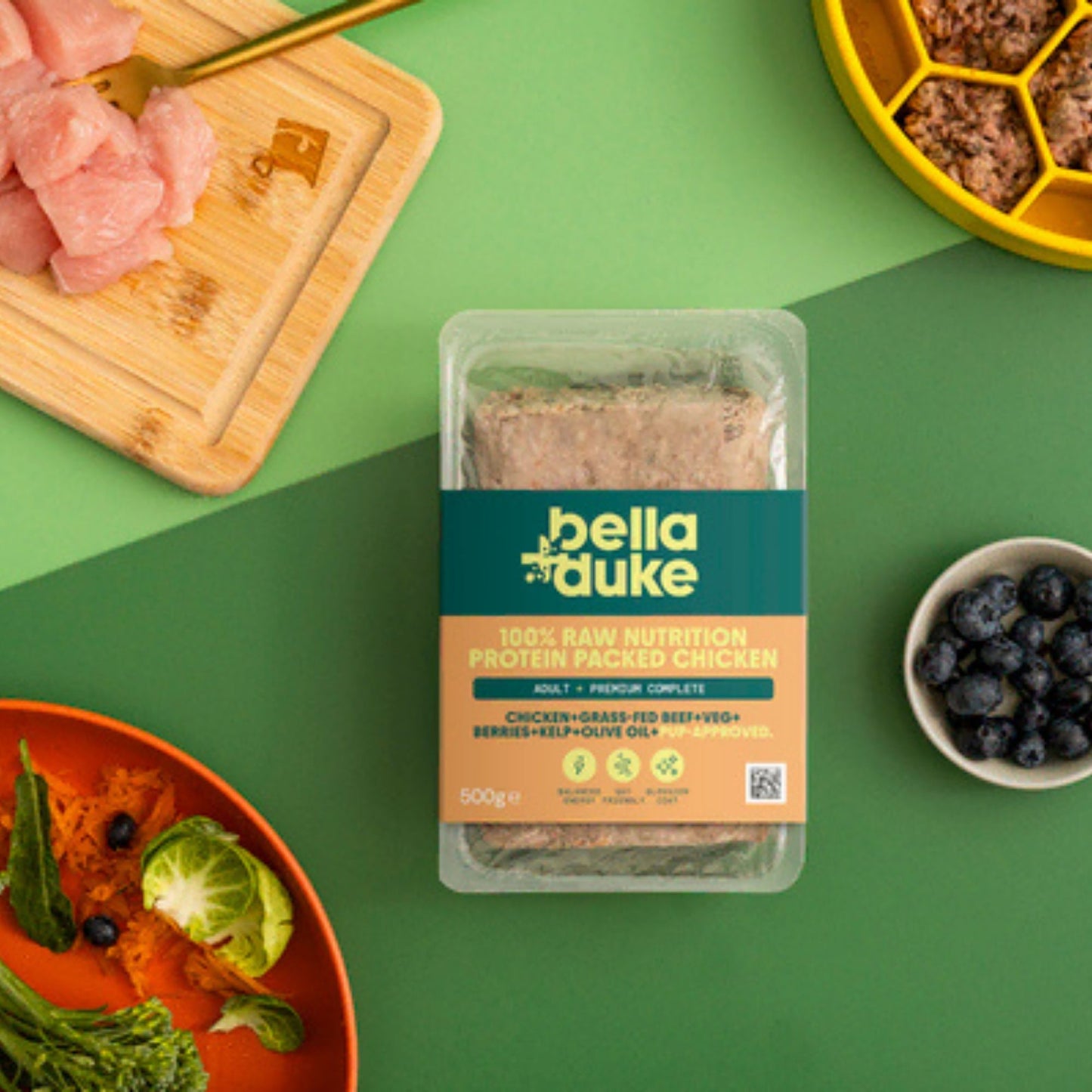 Bella+Duke pet food packaging on a green background with raw meat and vegetables.