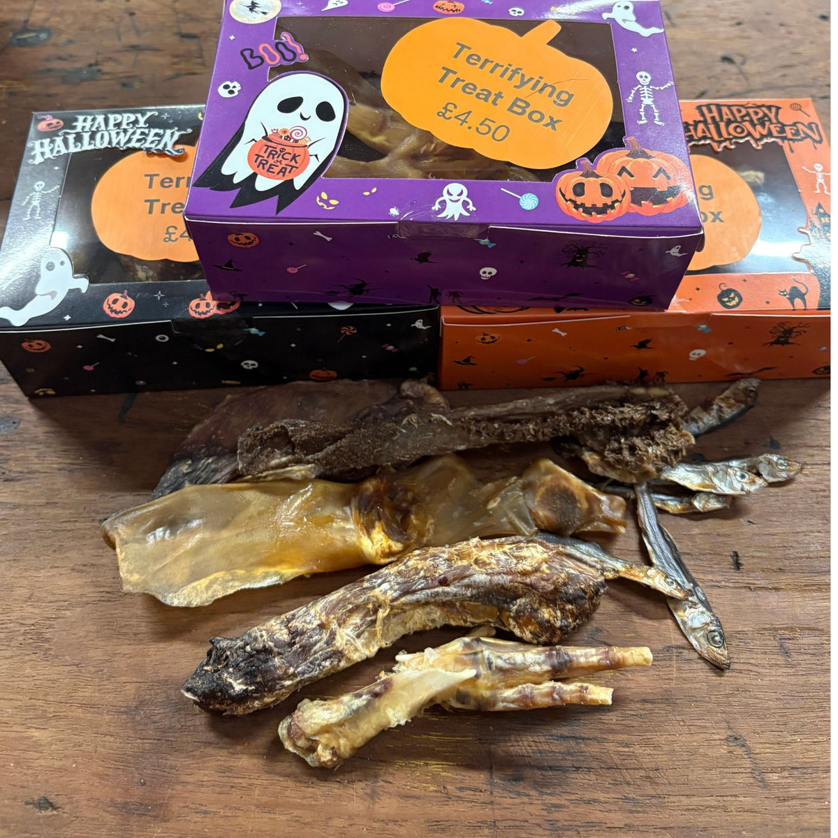 Three Halloween-themed treat boxes stacked on a wooden table, with dried animal jerky sticks and small dried fish pieces spread in front. The boxes are purple, black, and orange with 'Happy Halloween' and 'Terrifying Treat Box $4.
