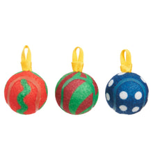 Three textured tennis balls shaped like Christmas ornaments hanging from yellow ribbons. The left ornament is red and green, the middle is green and red, and the right is blue with white polka dots.