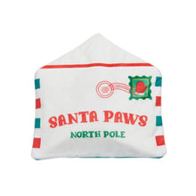 A white house-shaped plush dog toy designed to look like a Christmas letter envelope, featuring 'SANTA PAWS NORTH POLE' in red and green text, a red circular stamp graphic, and green and red striped borders on a white background.