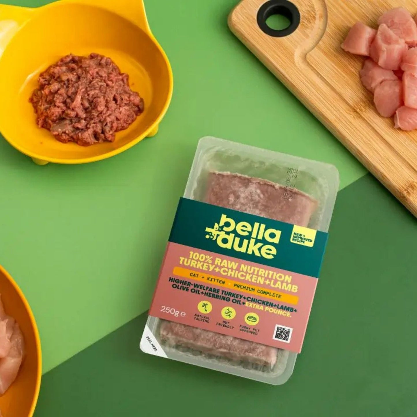 Packaged Bella+Duke pet food on a green background with raw meat and bowls.