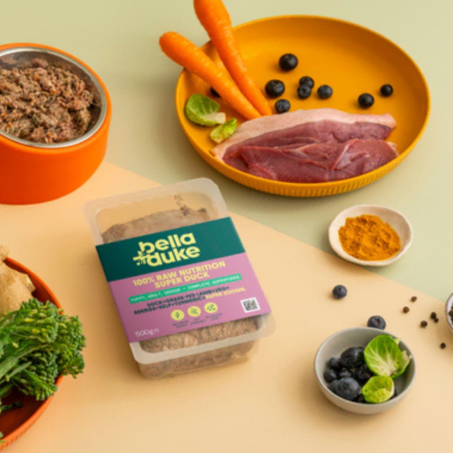 Bella+Duke pet food packaging with raw meat, vegetables, and a bowl of food on a light background