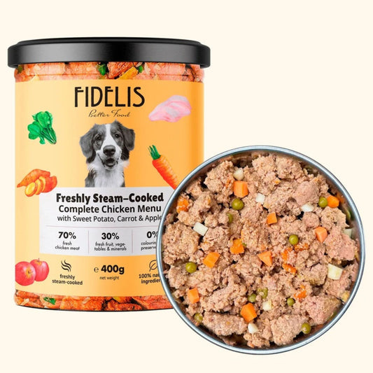 A container of FIDELIS Freshly Steam-Cooked Complete Chicken Meal wet dog food shown next to a metal bowl filled with the cooked chicken and vegetable food, on a light cream background.