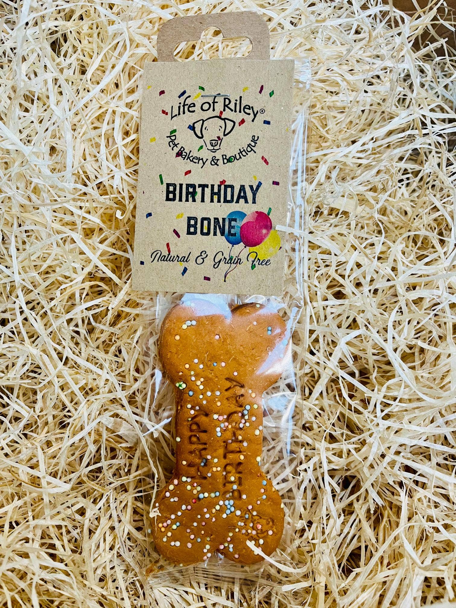 Dog bone-shaped treat with 'Happy Birthday' text on a straw background, branded 'Life of Riley'.