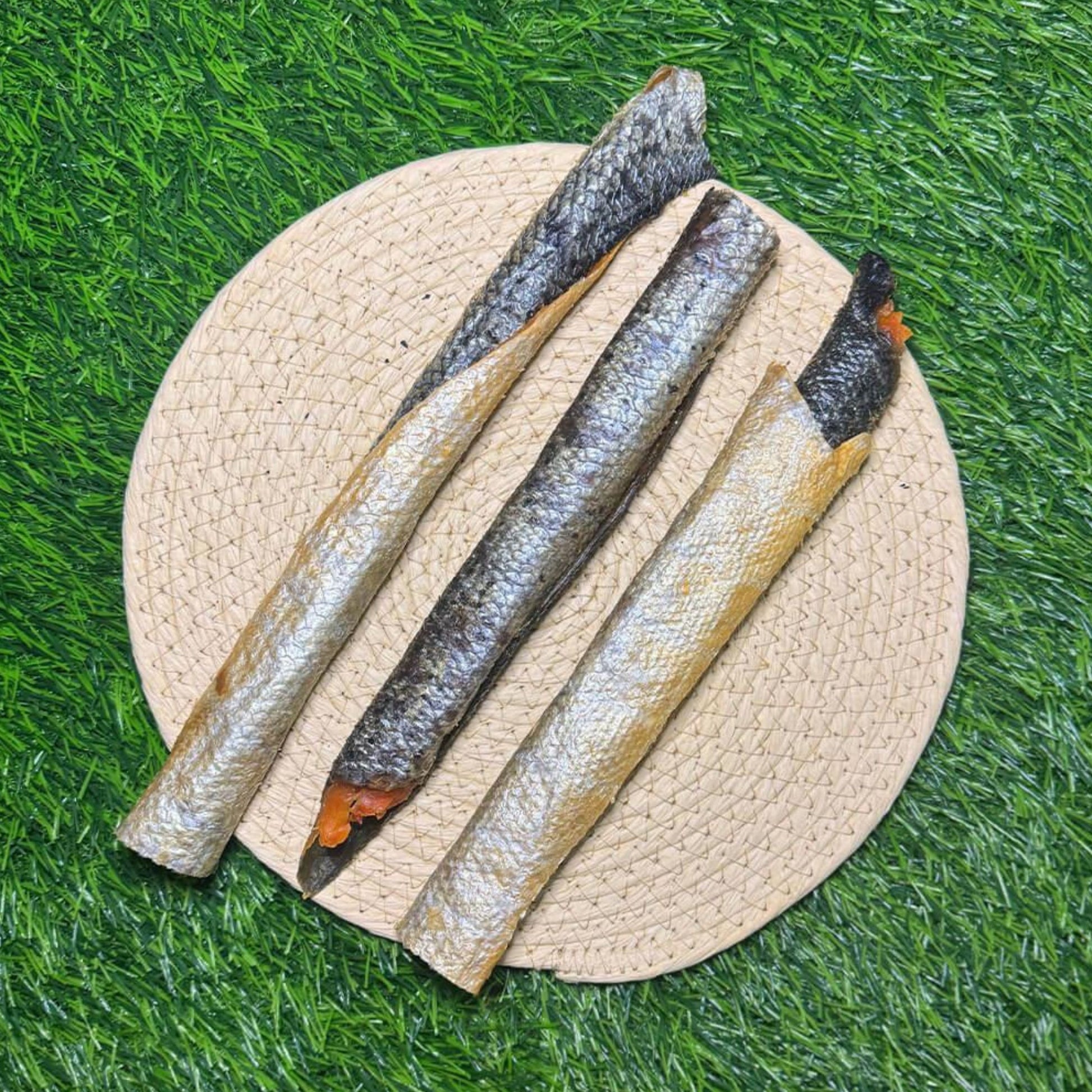 Three long, dried salmon arranged diagonally on a round woven straw mat, set against a bright green artificial grass background.
