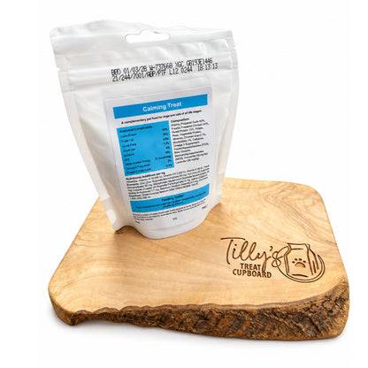 A white stand-up pouch labeled 'Calming Treat' with a blue nutrition panel, displayed on a wooden bark-edge tray engraved with 'Tilly's Treat Cupboard'.