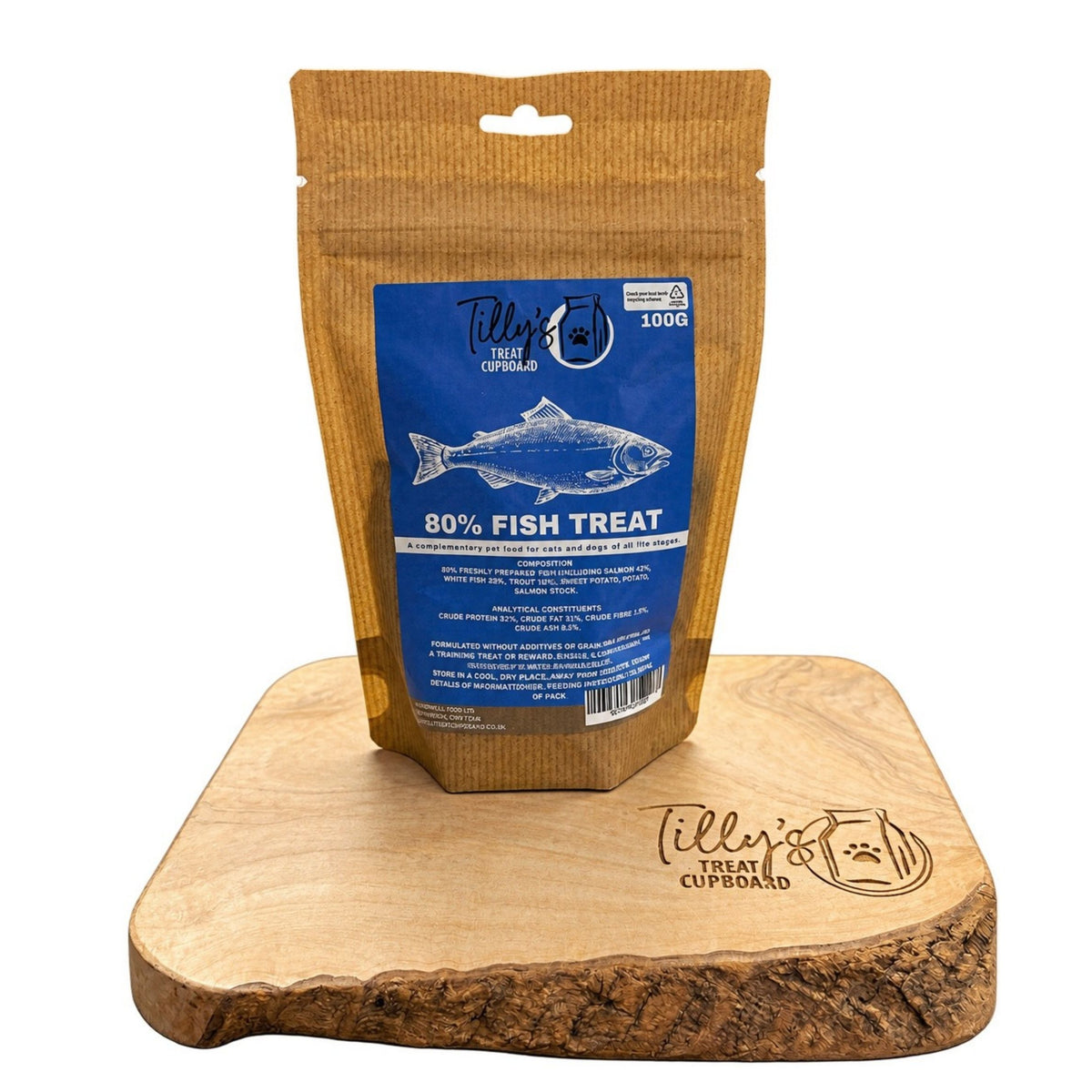 A 100g pouch of Tilly's Fish 80% Fish Treat for dogs, displayed on a wooden treat cupboard. The pouch is brown kraft paper with a blue label featuring a fish graphic.