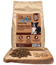 A bag of Tilly's Treats Cupcake Adult Dog grain-free dog food stands on a wooden cutting board with kibble pieces scattered in front. The bag is kraft brown with a large tan label featuring a black-and-white dog photo and product text.