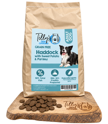 A bag of Tilly's Treat Cupboards Grain Free Haddock with Sweet Potato & Parsley dog food is shown on a wooden board with loose kibble pieces in front. The bag is kraft brown with a light blue label featuring a photo of a black and white dog.