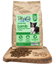 A bag of Tilly's Treat Cupboard Adult Dog Lamb with Sweet Potato & Mint dry dog food is displayed on a wooden cutting board with loose kibble pieces in front. The bag is kraft brown with a light green and white label featuring a black and white border collie.