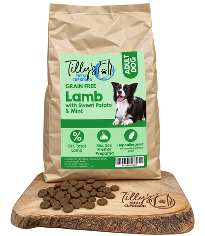 A bag of Tilly's Treat Cupboard Adult Dog Lamb with Sweet Potato & Mint dry dog food is displayed on a wooden cutting board with loose kibble pieces in front. The bag is kraft brown with a light green and white label featuring a black and white border collie.