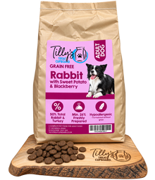A bag of Tilly's Treat Cupboard Adult Dog Rabbit food stands upright on a wooden cutting board with scattered brown kibble pellets in front. The bag is kraft paper with a large pink label showing a black-and-white dog photo and product text.