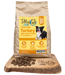 A bag of Tilly's Treat Cupboard adult dog food stands on a wooden board with scattered kibble pieces in front. The bag is kraft paper with a large yellow label reading 'Turkey with Sweet Potato & Cranberry' and showing a black-and-white dog photo.
