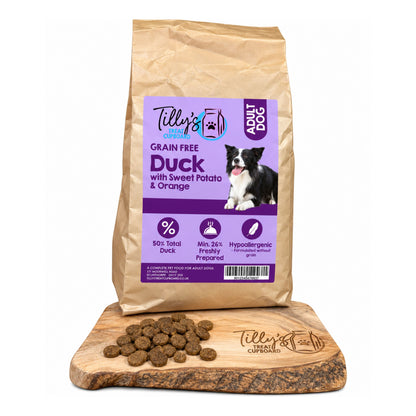 A bag of Tilly's Treat Cupboard Grain Free Duck with Sweet Potato & Orange dog food stands upright on a wooden board with some kibble spilled in front, against a plain white background.