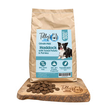 A bag of Tilly's Treat Cupboard Adult Dog food stands on a wooden board with loose kibble pieces in front. The package is kraft paper with a large light blue label showing the brand name, 'Haddock with Sweet Potato & Parsley' flavour, and a photo of a black-and-white dog.