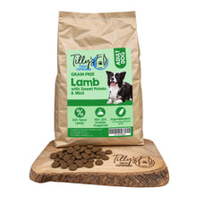 A bag of Tilly's Treat Cupboard Grain Free Lamb dog food with Sweet Potato & Mint, displayed on a wooden cutting board with kibble scattered in front. The bag is kraft brown with a light green and white label featuring a black and white dog.