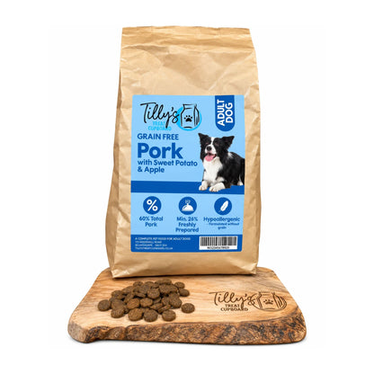 A bag of Telly's Grain Free Pork with Sweet Potato & Apple dog food stands upright on a wooden cutting board, with several brown kibble pieces scattered in front. The bag is kraft brown with a large blue front label and a photo of a black-and-white dog.