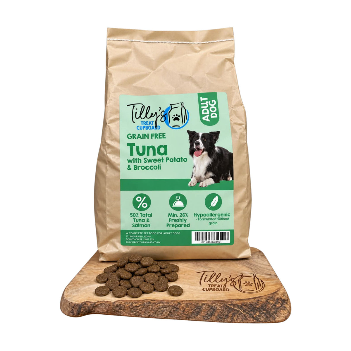 A bag of Tilly's Treats for Dogs Grain Free Tuna with Sweet Potato & Broccoli dog food stands upright on a wooden cutting board, with several brown kibble pieces scattered in front. The bag is kraft paper with a large green and white front label showing a dog photo and product details.
