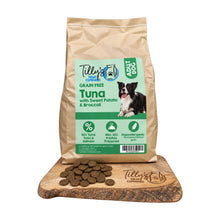 A bag of Tilly's Treats for Dogs Grain Free Tuna with Sweet Potato & Broccoli dog food stands upright on a wooden cutting board, with several brown kibble pieces scattered in front. The bag is kraft paper with a large green and white front label showing a dog photo and product details.