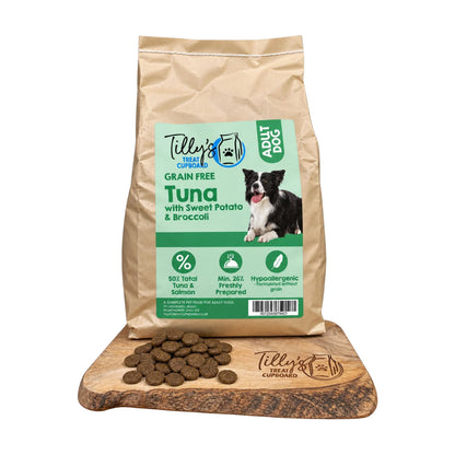 A bag of Tilly's Treats for Dogs Grain Free Tuna with Sweet Potato & Broccoli dog food stands upright on a wooden cutting board, with several brown kibble pieces scattered in front. The bag is kraft paper with a large green and white front label showing a dog photo and product details.