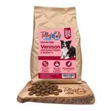 A bag of Tilly's Treat Cupboard Adult Dog grain-free venison kibble stands upright on a wooden board with loose kibble pieces in front. The package is kraft paper with a large pink front label showing the brand, product type, and a dog photo.