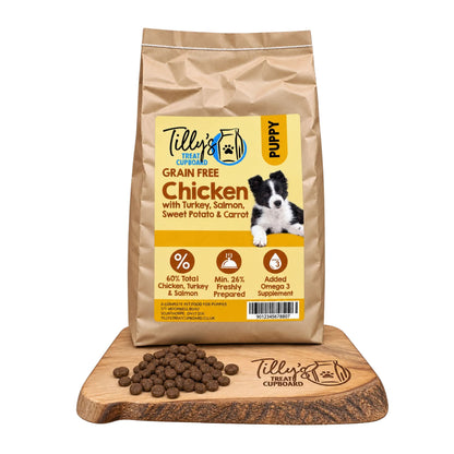 A bag of Tilly's Treat Cupboard Puppy Grain Free Chicken wet food stands on a wooden cutting board with loose kibble pieces in front. The bag is tan with a large yellow label and a photo of a black-and-white puppy.