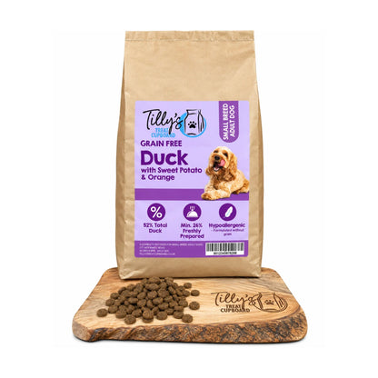 A bag of Tilly's Treat Cupboard Duck with Sweet Potato & Orange grain free pet food stands on a wooden cutting board with kibble scattered in front. The bag is kraft paper with a purple and white label, and the kibble is brown.