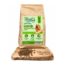 A bag of Tilly's Treat Cupboards Grain Free Lamb dog food stands upright on a wooden cutting board with loose kibble scattered in front. The package is kraft brown with a large green label, and the dog photo on the label shows a golden retriever.