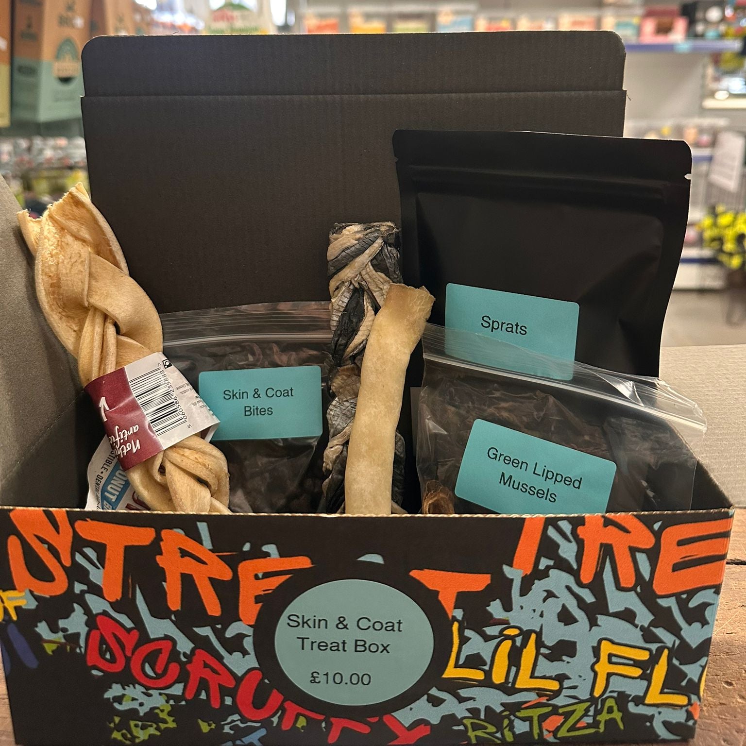 Dog treat box with various treats and packaging labeled 'Sprats', 'Skin & Coat Bites', and 'Green Lipped Mussels' in a store setting.