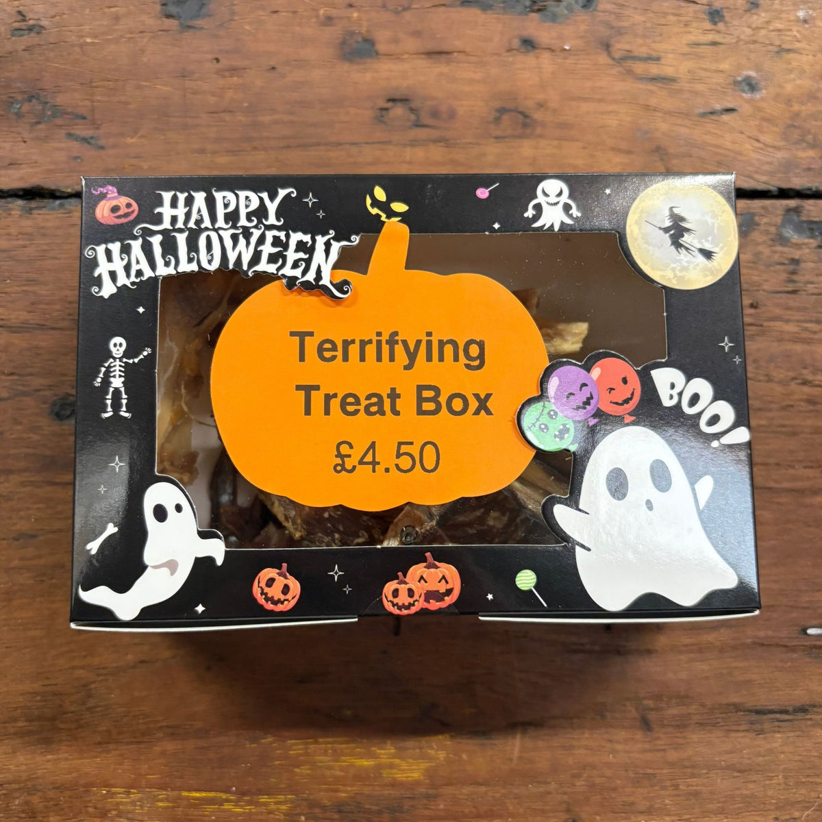 A Halloween-themed treat box on a rustic wooden table. The black box features 'Happy Halloween' text, ghost illustrations, jack-o'-lanterns, and a large orange label in the center that reads 'Terrifying Treat Box £4.