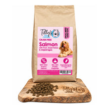 A bag of Tilly's Treat Cupboard Grain Free Salmon dog food stands upright on a wooden cutting board with loose kibble scattered in front. The packaging is kraft paper with a large pink label, and the dog pictured on the label is small and not the actual subject.
