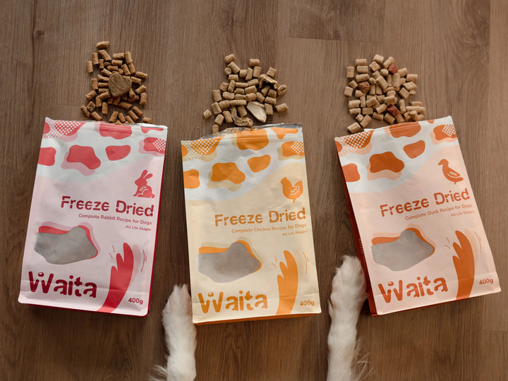 Three packages of Waita freeze-dried pet food on a wooden surface with pet treats above them.